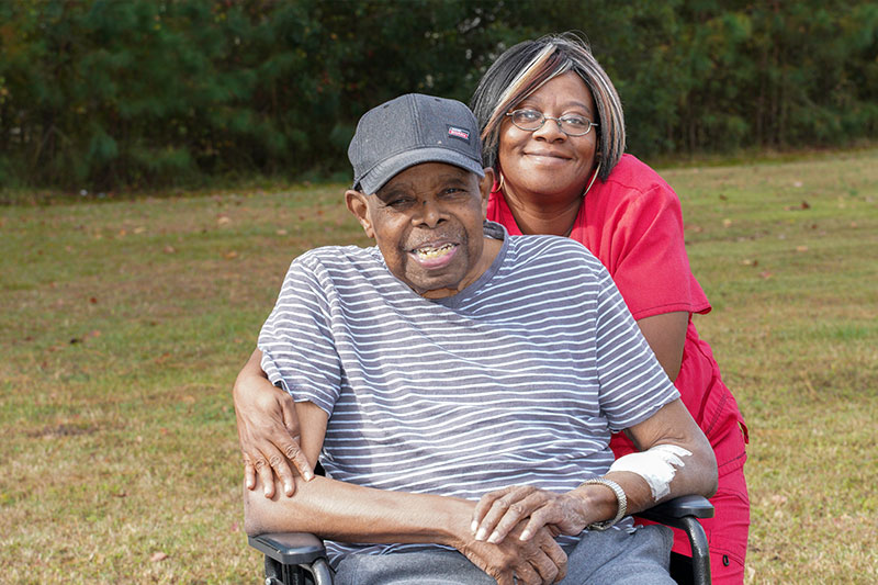 An Older African American adult sitting in a wheelchair with an East Carolina rehab staff member standing behind them