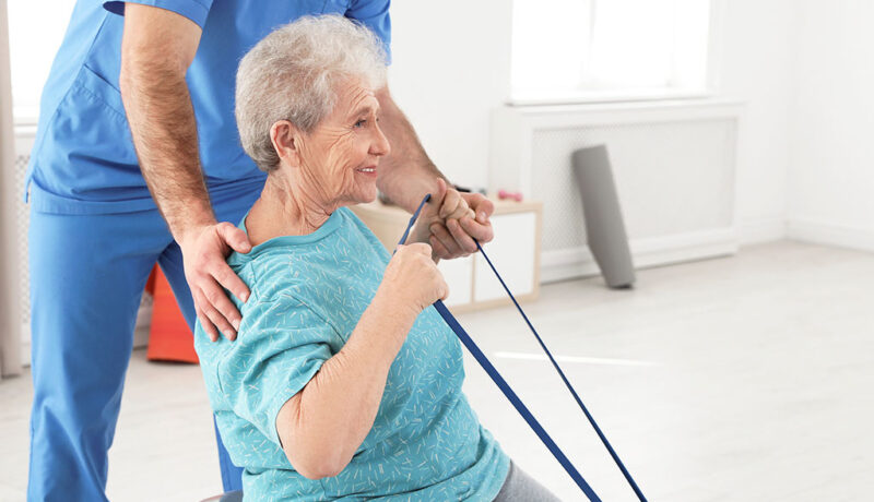 An Elderly Woman Sitting On A Yoga Ball Using A Resistant Band To Stretch With A Physical Therapist Assisting Her Short Vs Long-Term Physical Therapy