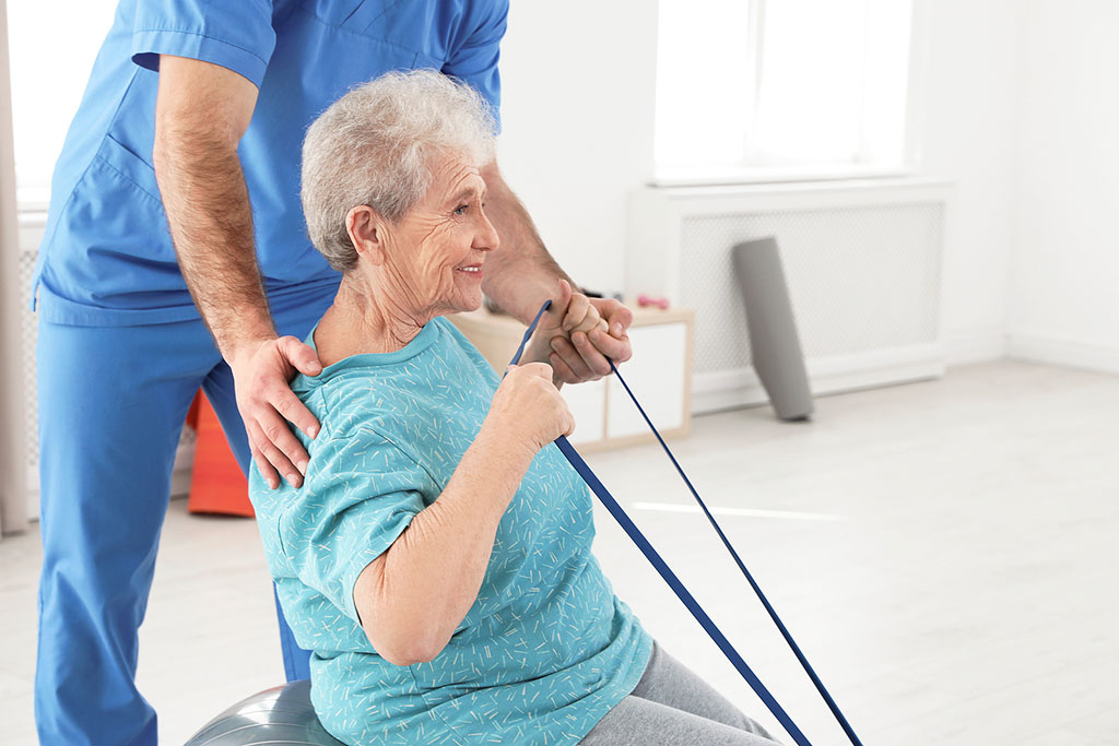 An Elderly Woman Sitting On A Yoga Ball Using A Resistant Band To Stretch With A Physical Therapist Assisting Her Short Vs Long-Term Physical Therapy